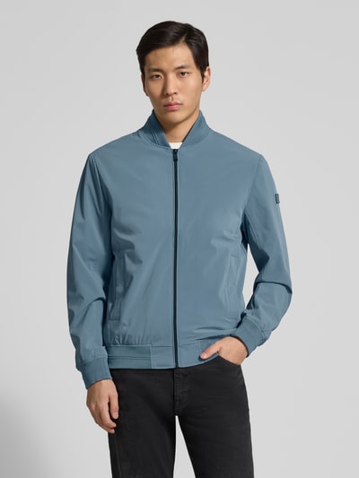 Mannenmodel draagt Regular Fit Bomberjacke mit Stehkragen van Guess in lichtblauw met zijzakken en elastische boorden, frontaal totaalbeeld.