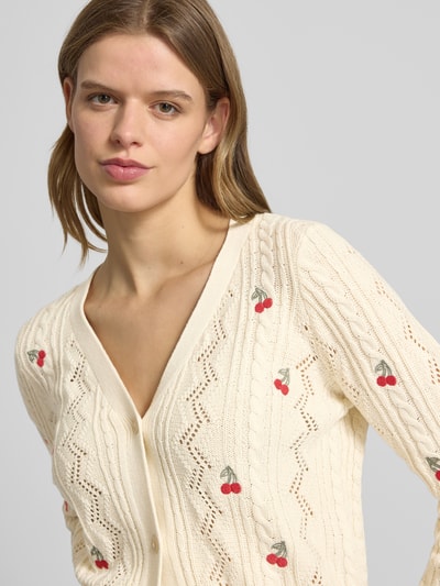 Modelka kobieca ma na sobie Regular Fit Strickjacke aus Baumwoll-Mix Modell 'BIRTIE' od Only, efekt: jednolity kolor, krój regular fit, listwa guzikowa, wzór: faktura, ścieg warkoczowy, aplikacja: hafty, kształt kołnierza: crew neck, dekolt: głęboki dekolt w serek, rękaw: długi, prążkowane zakończenia, wykończenie materiału: dzianina, materiał: mieszanka bawełny, jasny kremowy kolor, czerwone haftowane wisienki, fakturowany wzór, zbliżenie na górną część ciała, perspektywa z przodu.