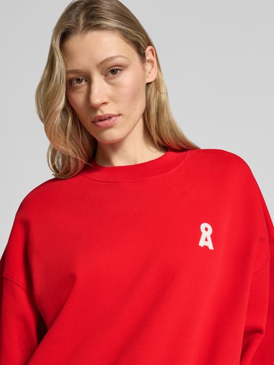 Frauenmodel trägt Oversized Sweatshirt aus reiner Baumwolle Modell 'ICONIC A ALIZAA' von Armedangels in Uni-Rot mit Logo-Stitching, Nahaufnahme Oberkörper.