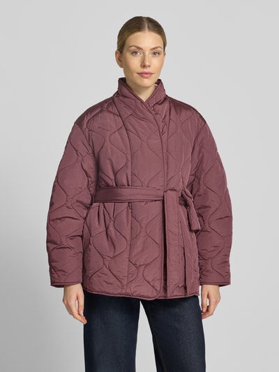 Modelka kobieca nosi Steppjacke mit Bindegürtel Modell 'Himiria' od YAS, krój regular fit, stójka, śliwkowy kolor, tekstura pikowana, pasek, Szerokość w rozmiarze 36: 55 cm, Długość pleców: 76 cm, Szerokość ramion: 49 cm, Długość rękawa: 57 cm, zdjęcie całej sylwetki z przodu.