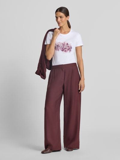 Kobiece model prezentuje Damski t-shirt Regular Fit T-Shirt aus reiner Baumwolle od Liu Jo White, biała bawełna, nadruk z logo, obszycie z kamieni stras, okrągły dekolt, długość tyłu przy rozmiarze S 66 cm, widok całej sylwetki z przodu.