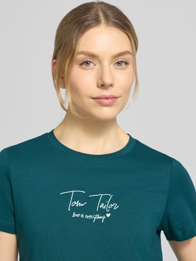 Kobiece model prezentuje Regular Fit T-Shirt aus Bio-Baumwoll-Mix von Tom Tailor, zielony jednolity, krótki rękaw, okrągły dekolt, napis, krój regular fit, materiał bio-bawełna, zbliżenie na górną część ciała