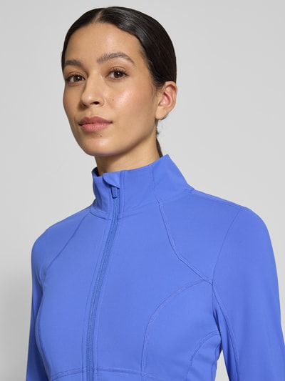 Frauenmodel trägt Slim Fit Jacke mit Stehkragen Modell 'Storm' von Fabletics in Blau, glatte Textur, Nahaufnahme Oberkörper.