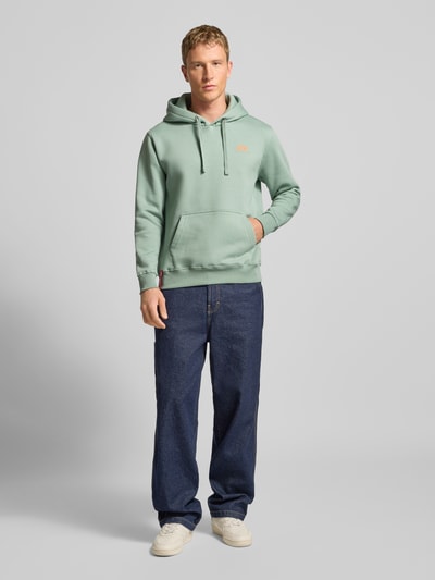 Męski model nosi Hoodie mit Logo und Känguru-Tasche od Alpha Industries, mieszanka bawełny, krój regular fit, dł. tyłu 69 cm, dł. rękawów 64 cm, jasny zielony, kieszeń kangurka, widok całej sylwetki z przodu.