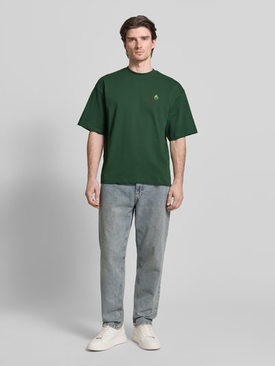 Męski model nosi T-Shirt mit Label-Detail Modell 'Beam' od WOODBIRD, zielona bawełna organiczna, luźny krój, widok całej sylwetki z przodu.