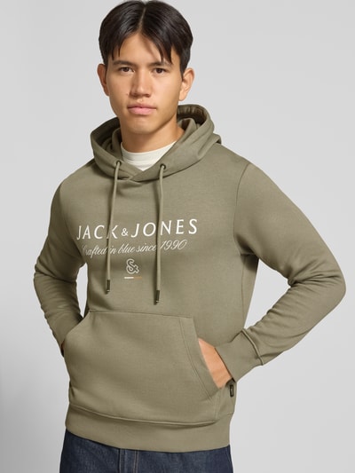 Männermodel trägt Relaxed Fit Hoodie aus Baumwoll-Mix Modell 'KYLE' von JACK & JONES in Olivgrün mit Känguru-Tasche, Ganzkörperansicht von vorne. Rückenlänge 67 cm, Ärmellänge 65 cm, Kapuze mit Tunnelzug, Label-Detail.