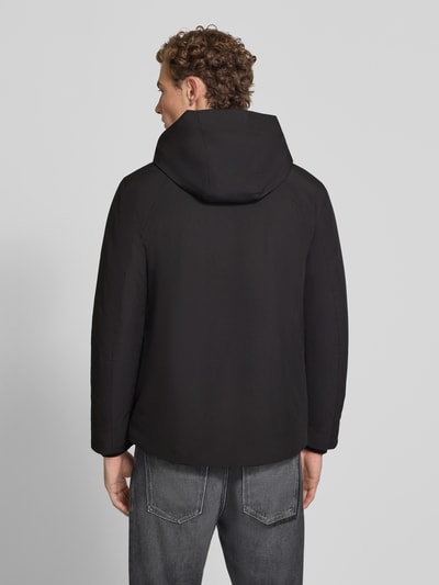 Męski model nosi Regular Fit Jacke mit Logo-Print von Lindbergh, czarna elastyczna mieszanka materiałów, kaptur z tunelem, nadruk z logo, napoleońska kieszeń, długość tyłu w rozmiarze M: 74 cm, szerokość ramion w rozmiarze M: 46 cm, szerokość klatki piersiowej w rozmiarze M: 42 cm, długość rękawów w rozmiarze M: 62 cm, widok całej sylwetki z tyłu.
