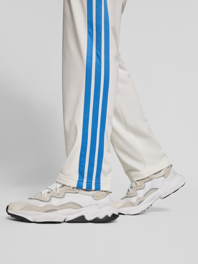 Männermodel trägt Straight Fit Sweatpants mit Logo-Stitching von adidas Sportswear in Weiß mit blauen Galonstreifen, Detailaufnahme. Herrenmodell mit elastischem Bund und Tunnelzug, Innenbeinlänge bei Größe M 80 cm.