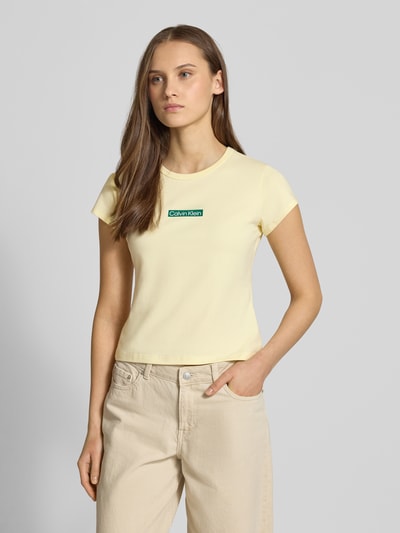 Modelka nosi T-shirt mit Rundhalsausschnitt und Label-Print von Calvin Klein Jeans, mieszanka bawełny i elastanu, krój slim fit, okrągły dekolt, nadruk z logo, długość tyłu w rozmiarze S: 42 cm, jasny kremowy kolor, widok całej sylwetki z przodu.