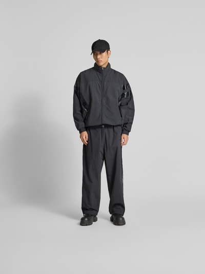 Männermodel trägt schwarze Trainingsjacke mit Stehkragen von VETEMENTS, Relaxed-Fit, Zweiwege-Reißverschluss, elastische Abschlüsse, Eingrifftaschen, Ganzkörperansicht von vorne. Rückenlänge 74 cm, Schulterbreite 50 cm, Ärmellänge 73 cm.