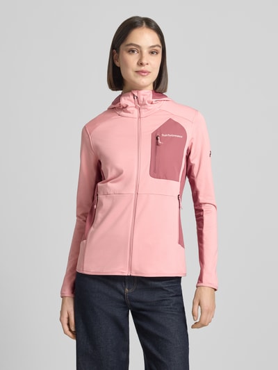 Frauenmodel trägt Jacke mit Logo-Stitching von PEAK PERFORMANCE in Rosa, Elastischer-Material-Mix, körpernahe Passform, Kapuze, Reißverschluss, Brusttasche mit Reißverschluss, Ganzkörperansicht von vorne.