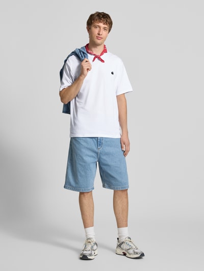 Männermodel trägt Herren T-Shirt mit Label-Stitching Modell 'Madison' von Carhartt Work In Progress, Reine Baumwolle, Regular-Fit, Rundhalsausschnitt, blaue Shorts, helle Sneaker, hellgrauer Hintergrund, Ganzkörperansicht von vorne.
