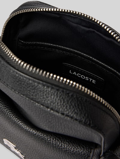 Produktfoto, Umhängetasche mit Label-Applikation von Lacoste aus schwarzem, genarbtem Leder, rechteckige Form, sichtbares Hauptfach mit Reißverschluss und Label, unifarbene Optik, Nahaufnahme.