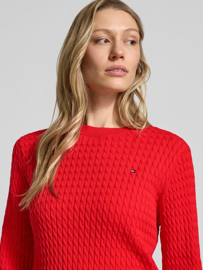 Frauenmodel trägt Slim Fit Pullover aus reiner Baumwolle von Tommy Hilfiger in Rot, Zopfmuster, Logo-Detail, Nahaufnahme Oberkörper.