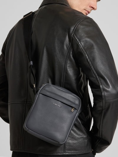 Männermodel trägt schwarze Lederjacke und Handtasche mit Label-Detail Modell 'RAISED REPORTER' von CK Calvin Klein, Ganzkörperansicht Rückansicht. Hauptfach mit Reißverschluss, Schulterriemen, strukturierte Oberfläche, kompaktes rechteckiges Design.