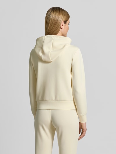 Modelka nosi Sweatjacke mit Label-Applikation Modell 'OLYMPE' od Guess Activewear, kolor jasny kremowy, luźny krój, miękka tekstura, kaptur, z mieszanki materiałów z dodatkiem modalu, widok całej sylwetki tyłem
