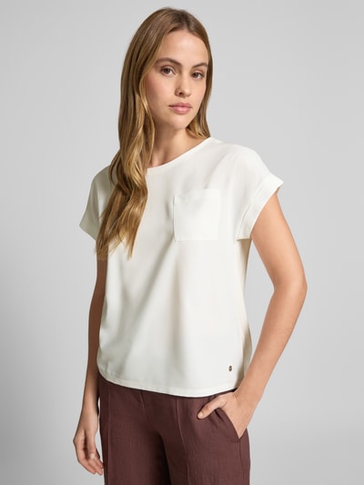 Frauenmodel trägt Blusenshirt mit verlängertem Rücken und Brusttasche von monari in Creme mit Gerader-Schnitt, Rundhalsausschnitt, Aufschlag an den Ärmeln, Brusttasche, Label-Detail, glatter Struktur, Rückenlänge 54 cm, Ärmellänge 3 cm, Ganzkörperansicht von vorne.