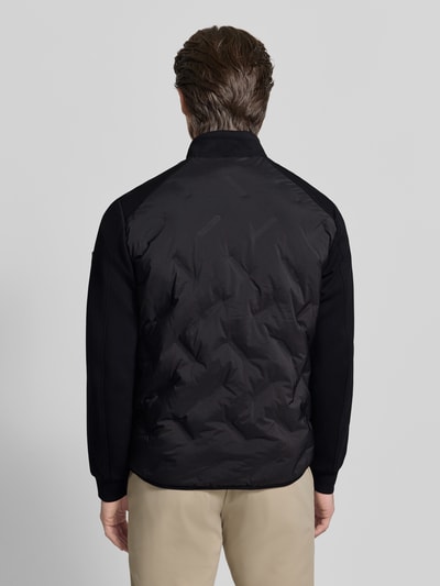 Männermodel trägt Regular Fit Jacke mit Stehkragen von Guess in Schwarz mit elastischem Material-Mix und Reißverschlusstaschen, Ganzkörperansicht Rückansicht.