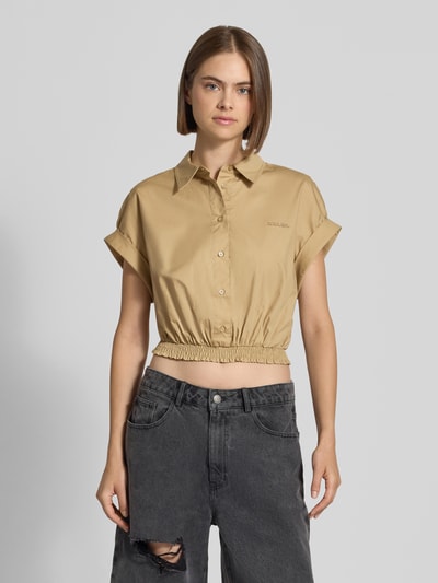 Modelka kobieca nosi Cropped Hemdbluse aus reiner Baumwolle mit Logo-Stitching von Karo Kauer w kolorze beżowym, krój krótki, wyhaftowane logo, kołnierzyk koszulowy, krótki rękaw, listwa guzikowa, elastyczny pas, długość tyłu w rozmiarze S: 45 cm, widok całej sylwetki z przodu.