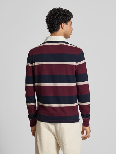 Männermodel trägt Regular Fit Rugby Shirt mit Logo- und Motiv-Stitching von Superdry in Bordeaux, Marine und Creme, Reine Baumwolle, Rückenlänge bei Größe M: 70 cm, Polokragen und kurze Knopfleiste, Label-Patch, Ganzkörperansicht Rückansicht.