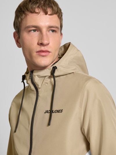 Männermodel trägt Jacke mit Kapuze und Ritssluiting Modell 'RUSH' von JACK & JONES in beige mit zwart logo, regular fit, steekzakken, detailaufnahme oberkörper. Ruglengte maat M: 68 cm, schouderbreedte: 46 cm, borstwijdte: 55 cm, mouwlengte: 68 cm.