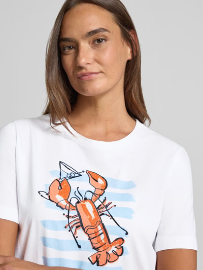 Kobiece modelka nosi T-Shirt mit Motiv-Print und Pailletten von Marc Cain z czystej bawełny, krój regular fit, okrągły dekolt, motyw nadruku z cekinami, kolor biały z pomarańczowym i niebieskim, długość tyłu w rozmiarze 36: 56 cm, zbliżenie na górną część ciała