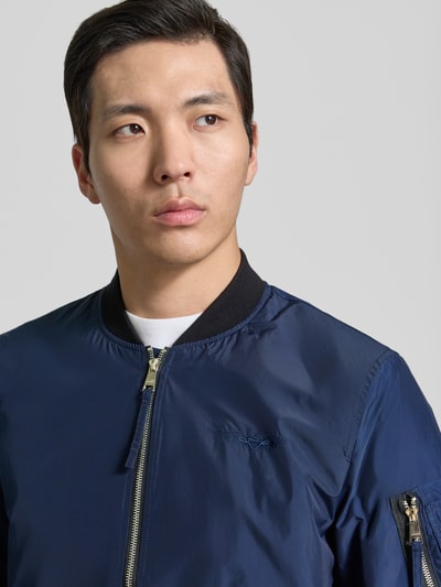Männermodel trägt Bomberjacke mit Label-Patch und Eingrifftaschen von Original Bombers in Dunkelblau, Nahaufnahme Oberkörper, prosty krój, zamek błyskawiczny, kieszeń na rękawie na zamek, kieszenie na zatrzask, ściągacze, długość tyłu w rozmiarze M: 67 cm, szerokość ramion w rozmiarze M: 43 cm, szerokość w rozmiarze M: 53 cm, długość rękawa w rozmiarze M: 68 cm