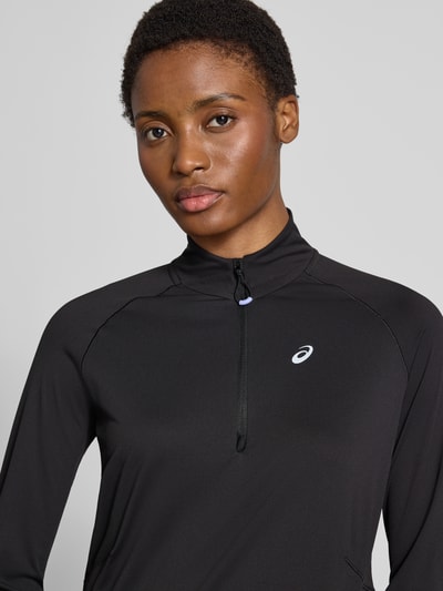 Frauenmodel trägt Longsleeve mit Troyer-Kragen Modell 'NAGINO' von ASICS TIGER in Schwarz mit reflektierendem Logo-Print, Regular Fit, Nahaufnahme Oberkörper.