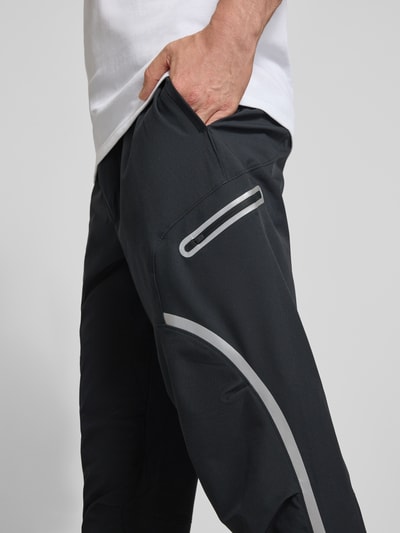 Männermodel trägt Regular Fit Sweatpants mit Logo-Print Modell 'Unstoppable' von Under Armour, Schwarz mit Cargo-Tasche, Nahaufnahme Oberschenkel. Elastischer-Material-Mix, Reißverschluss, Elastischer Bund, Innenbeinlänge 79 cm Größe M.