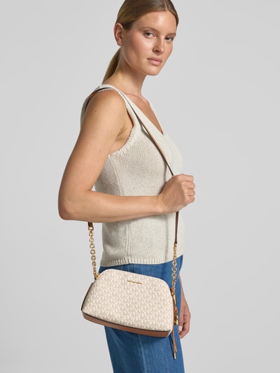 Frauenmodel trägt Handtasche mit Logo-Muster Modell 'FREDDIE' von MICHAEL Michael Kors, cremefarbene Tasche, goldfarbener Schulterriemen, Ganzkörperansicht von vorne.