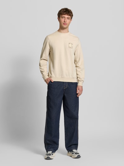 Männermodel trägt Regular Fit Sweatshirt mit floraler Applikation Modell 'Felica' von Blend in Beige, kombiniert mit Jeans und Sneakern, reine Baumwolle, Rundhalsausschnitt, elastische Abschlüsse, Ganzkörperansicht von vorne.