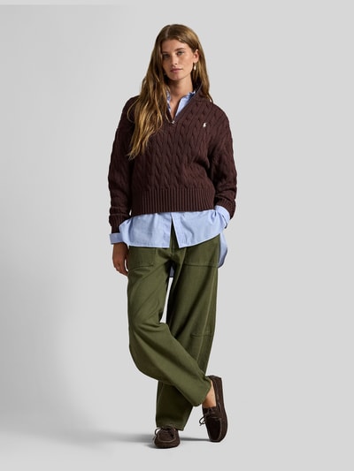 Frauenmodel trägt Troyer mit Zopfstrickmuster von Polo Ralph Lauren in Dunkelbraun, kombiniert mit Hemd und grüner Hose, Ganzkörperansicht von vorne. Modell besteht aus reiner Baumwolle, Regular Fit, Stehkragen mit Reißverschluss, Logo-Stitching, elastische Abschlüsse.