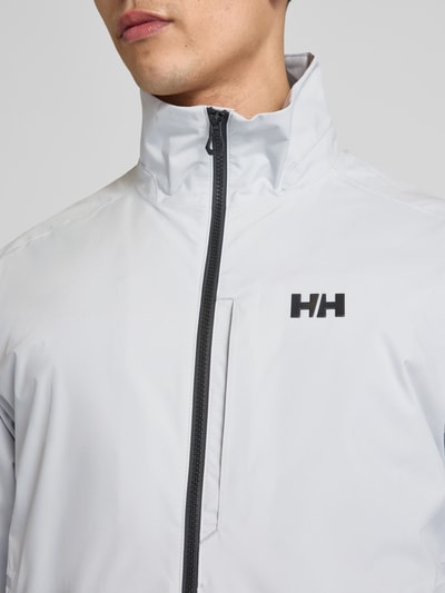 Männermodel trägt Bomberjacke mit Stehkragen Modell 'RACING' von Helly Hansen, Logo-Print, Reißverschluss, Nahaufnahme Oberkörper.