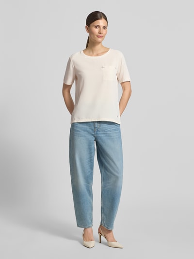 Kobiece modelka nosi damski t-shirt 'T-Shirt aus Viskose-Mix mit Brusttasche' od monari, mieszanka wiskozy z dodatkiem streczu, krój loose fit, okrągły dekolt, brustasza, obszycie z kamieni stras, detal z logo, długość tyłu w rozmiarze 36: 56 cm, jasny kolor, klasyczny fason, gładka tekstura, całkowity widok z przodu.