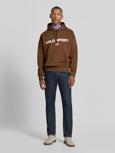 Mannenmodel draagt Relaxed Fit Hoodie aus Baumwoll-Mix van Polo Ralph Lauren in bruin met witte letters, blauwe jeans, beige sneakers, Ganzkörperaanzicht van voren.