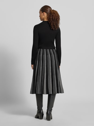 Modelka nosi Midikleid aus Viskose-Mix von Apricot, czarna góra, wzorzysta czarno-biała spódnica, imitacja dzianiny, stójka, taliowany krój, długi rękaw, długość tyłu w rozmiarze S: 108 cm, szerokość klatki piersiowej w rozmiarze S: 37 cm, długość rękawów w rozmiarze S: 61 cm, widok całej sylwetki z tyłu.
