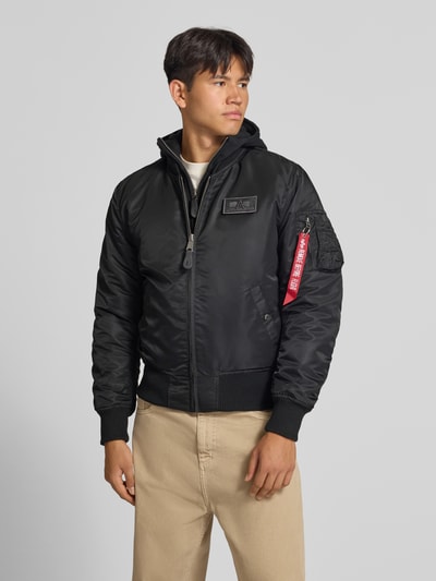 Männermodel trägt Jacke mit Reißverschlusstasche am Ärmel von Alpha Industries in Schwarz, Label-Patch, gerippte Abschlüsse, Ganzkörperansicht von vorne. Rückenlänge 64 cm, Schulterbreite 54 cm, Brustweite 54 cm, Ärmellänge 72 cm, Regular Fit.