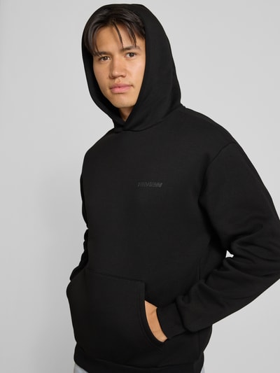 Männermodel trägt Relaxed Fit Sweatshirt aus Baumwoll-Mix mit Känguru-Tasche von REVIEW in Schwarz, sichtbares Label-Detail, Nahaufnahme Oberkörper.