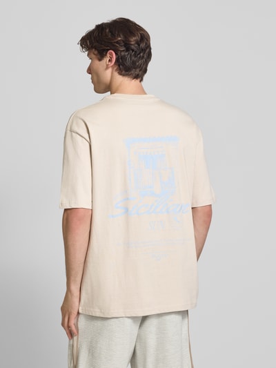 Männermodel trägt męski t-shirt Oversized T-Shirt mit rückseitigem Print Modell 'Pedroche' von Pegador, beżowa bawełna, krój oversized fit, nadruk z motywem i logo z tyłu, okrągły dekolt, długość tyłu w rozmiarze M: 71 cm, Ganzkörperansicht Rückansicht.