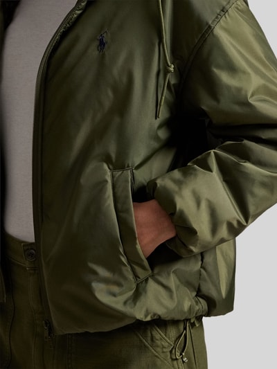 Kobieta nosi Relaxed Fit Blouson mit Zweiwege-Reißverschluss od Polo Ralph Lauren w kolorze khaki, jednolity, z logo na piersi, dwukierunkowy zamek, kaptur z troczkiem, elastyczne mankiety i dół, boczne kieszenie, detale z połyskiem, zbliżenie na górną część ciała.