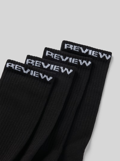 Produktfoto, schwarze Socken aus Baumwoll-Mix im 4er-Pack von REVIEW mit weißem Logo-Detail und elastischem Bund, neutraler Hintergrund.