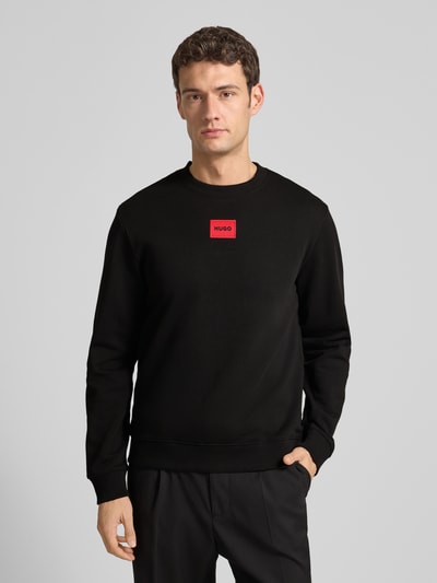 Männermodel trägt Sweatshirt aus Baumwolle Modell 'Diragol' von HUGO in Schwarz mit rotem Label-Patch, Gerader-Schnitt, Ganzkörperansicht von vorne.