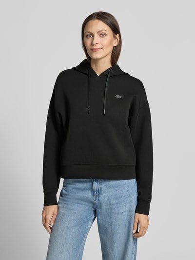 Frauenmodel trägt Oversized Hoodie aus Baumwoll-Mix von Lacoste Sport in Schwarz, langarm, Kapuze mit Tunnelzug, Logo-Detail, Nahaufnahme Oberkörper.