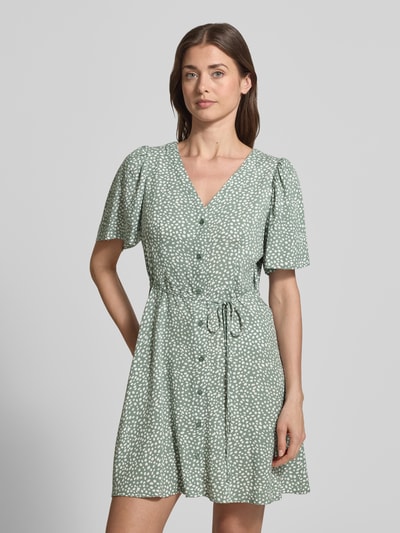 Modelka kobieca nosi Leicht ausgestelltes Minikleid aus reiner Viskose Modell 'ALBA' od Vero Moda, gładki wzór, mini długość, krótki szeroki rękaw, V-dekolt, pasek w talii, czysta wiskoza, kolor zielony z białymi kropkami, widok całej sylwetki z przodu.