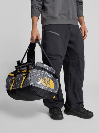 Männermodel trägt Weekender mit Logo-Print von The North Face in Schwarz mit gelben Details, geräumiges Hauptfach, Logo seitlich, Ganzkörperansicht von vorne.