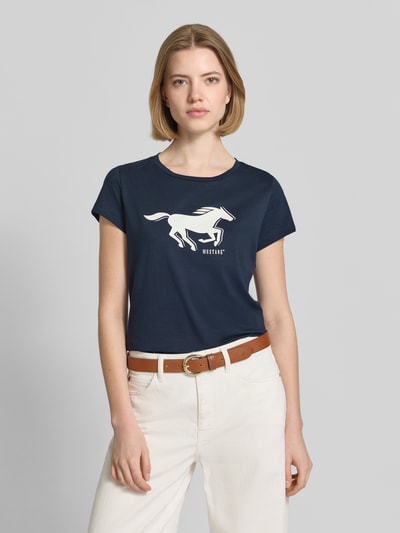 Kobiece model nosi damski t-shirt z bawełny z nadrukiem z logo, krój regular fit, model 'Loa' od Mustang, okrągły dekolt, ciemnoniebieski, długość tyłu w rozmiarze S: 60 cm, widok całej sylwetki z przodu.