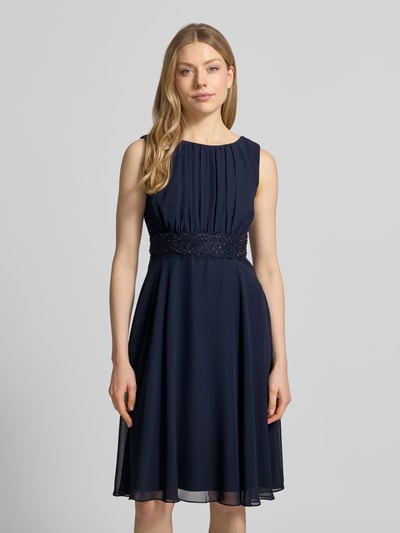 Frauenmodel trägt Knielanges Cocktailkleid mit Taillenband von Christian Berg Cocktail aus dunkelblauem Chiffon mit Drapierungen und Rundhalsausschnitt, taillierter Schnitt, Knielänge, Rückenlänge bei Größe 36: 103 cm, Brustweite bei Größe 36: 36 cm, Ganzkörperansicht von vorne.