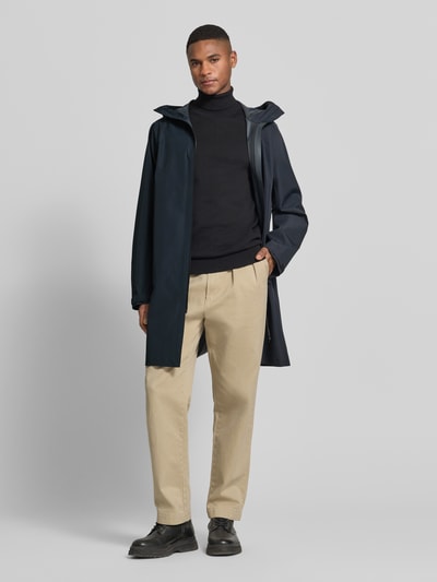Männermodel trägt Regular Fit Mantel mit Logo-Detail Modell 'Treeline' von Peak Performance in Dunkelblau, mit Kapuze, Zweiwege-Reißverschluss und Eingrifftaschen, kombiniert mit schwarzem Pullover und beiger Hose, Ganzkörperansicht von vorne.