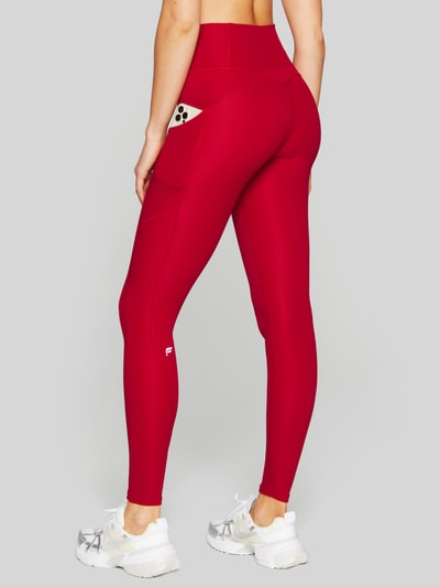 Frauenmodel trägt Slim Fit Leggins mit Beintaschen Modell 'OASIS' von Fabletics in Rot, elastischer Material-Mix, Ganzkörperansicht Rückansicht.