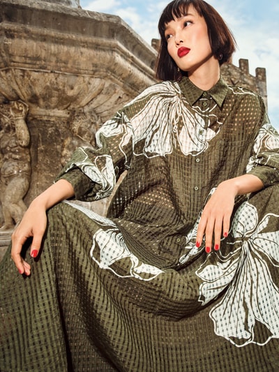 Frauenmodel trägt A-Linien Midi-Rock mit Kellerfalten am Bund Modell 'JANGO' von Essentiel Antwerp, florales Allover-Muster, transparenter Besatz, blickdichter Unterrock, Motiv-Print, Eingrifftasche, Midi-Länge, olivegrüne und weiße Farbtöne, strukturierte Textur, Ganzkörperansicht von vorne.